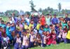anak-anak pemain sepak bola usia dini bersama pelatih dan pejabat daerah berpose sambil mengangkat tangan di atas lapangan hijau.