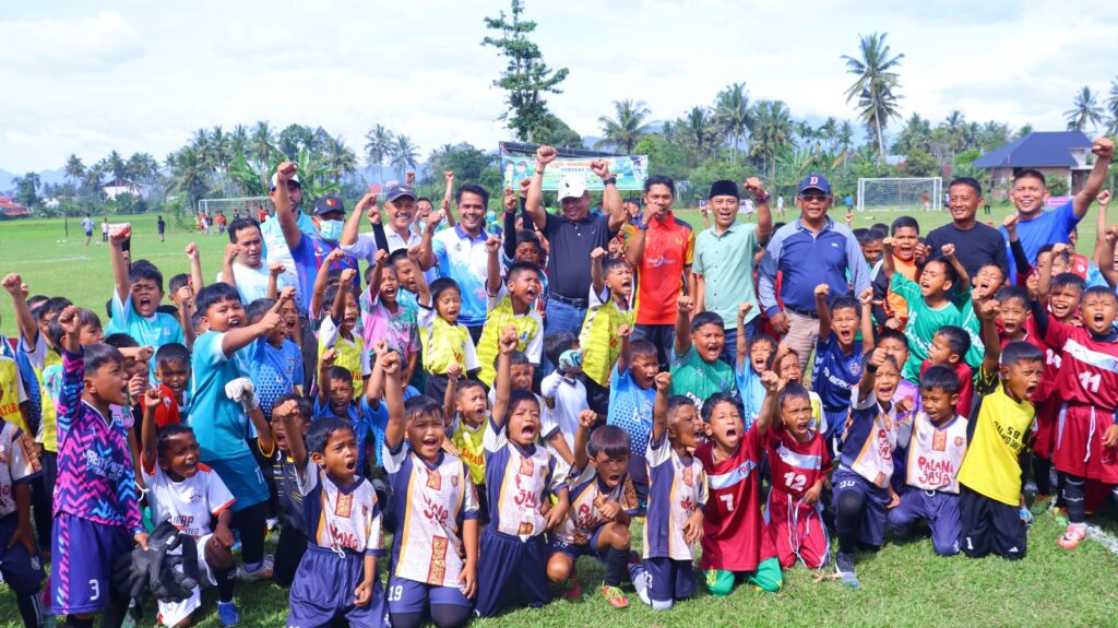 anak-anak pemain sepak bola usia dini bersama pelatih dan pejabat daerah berpose sambil mengangkat tangan di atas lapangan hijau.