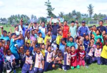 anak-anak pemain sepak bola usia dini bersama pelatih dan pejabat daerah berpose sambil mengangkat tangan di atas lapangan hijau.