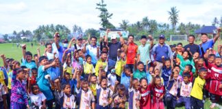 anak-anak pemain sepak bola usia dini bersama pelatih dan pejabat daerah berpose sambil mengangkat tangan di atas lapangan hijau.