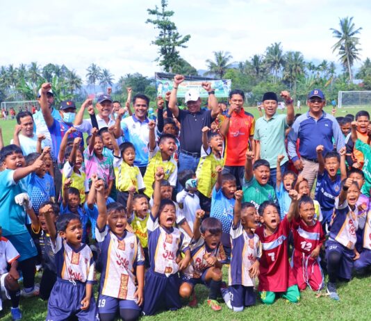 anak-anak pemain sepak bola usia dini bersama pelatih dan pejabat daerah berpose sambil mengangkat tangan di atas lapangan hijau.
